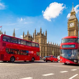 Deux bus rouges à impériale sur une rue avec les Chambres du Parlement et Big Ben en arrière-plan sous un ciel bleu.