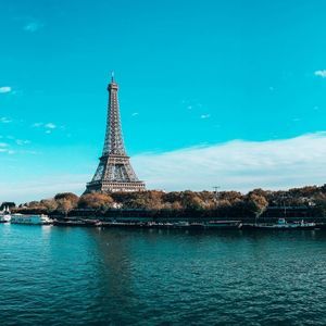 The Eiffel Tower stands across a wide river lined with autumn trees and moored boats, under a bright blue sky.