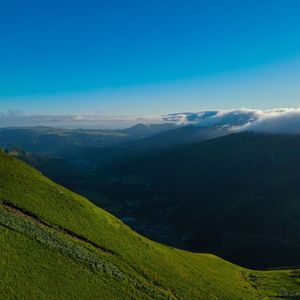 Une pente de montagne raide et herbeuse surplombe une vallée ombragée avec des nuages recouvrant les sommets lointains sous un ciel bleu éclatant.