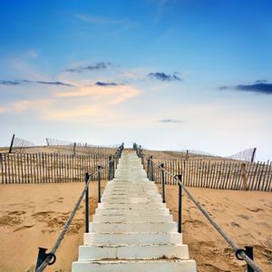 Un escalier blanc avec des rampes en corde monte une dune de sable, menant vers un ciel bleu parsemé de nuages.