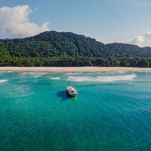 Veduta aerea di una barca a vela bianca ancorata in acqua turchese, con una spiaggia sabbiosa e una montagna verde lussureggiante sullo sfondo.