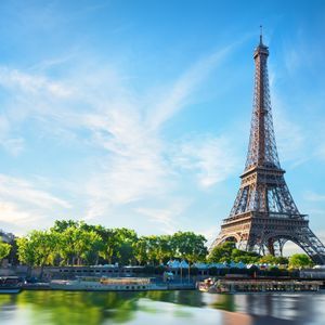 The Eiffel Tower stands over the Seine River with boats docked along the tree-lined bank under a clear blue sky.