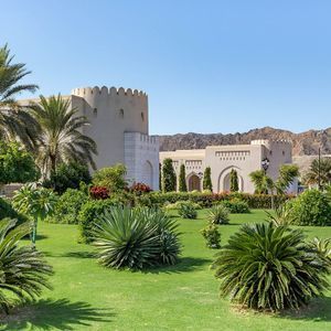 Un rigoglioso giardino verde pieno di palme e piante di fronte a un edificio tradizionale, con montagne rocciose sotto un cielo sereno.