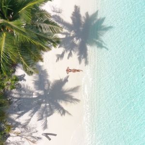 Vista aérea de una mujer relajándose en una playa de arena blanca, con sombras de palmeras y agua turquesa cristalina.