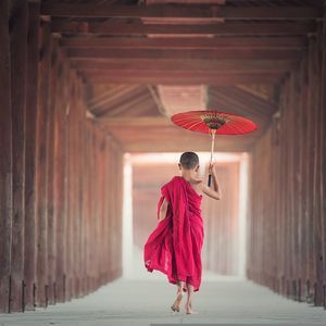 Un joven monje con túnicas rojas camina descalzo por un largo pasillo bordeado de pilares de madera, sosteniendo un paraguas rojo.