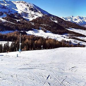 Vista panoramica di un'ampia pista da sci battuta in una giornata di sole, con montagne e foreste innevate sullo sfondo.