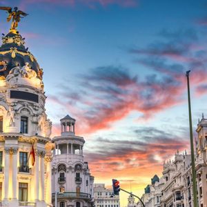 Edifici cittadini decorati, uno con tetto a cupola e statua dorata, sono illuminati contro un cielo vibrante al tramonto.