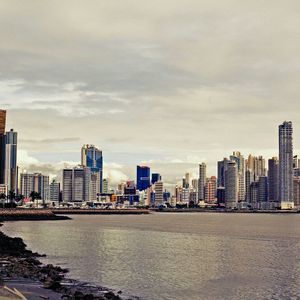 Skyline moderno di città con numerosi grattacieli, visto da una costa rocciosa oltre l'acqua sotto un cielo nuvoloso.