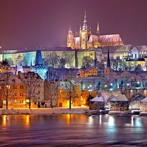Una città innevata con un grande castello e un ponte di pietra, illuminata di notte con luci che si riflettono nel fiume.