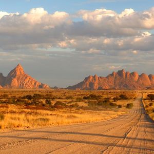 Una lunga strada sterrata si estende attraverso una secca pianura della savana verso distanti montagne rocciose illuminate dal sole sotto un cielo parzialmente nuvoloso.