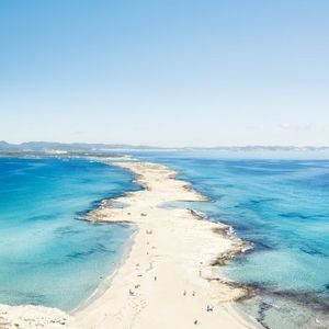 Vue aérienne d'une étroite langue de sable s'étendant dans une mer turquoise, avec un voilier ancré au large sous un ciel bleu clair.