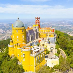 Una vista aerea di un grande castello colorato con torri gialle e rosse, situato su una collina verde e alberata sotto un cielo azzurro e limpido.