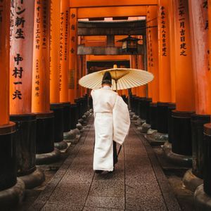 Una persona in abiti tradizionali bianchi cammina con un ombrello di carta lungo un sentiero fiancheggiato da cancelli torii arancioni.