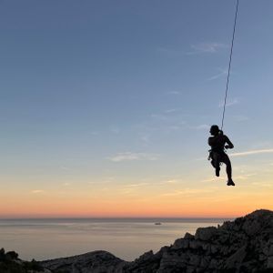 Une personne en silhouette dans un harnais, suspendue à une corde au-dessus de falaises rocheuses et de la mer au coucher du soleil.