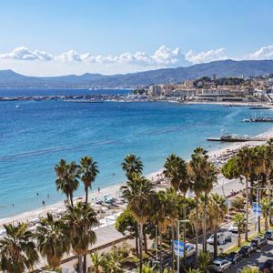 An aerial view of a coastal city with a bustling sandy beach, blue sea, and mountains in the background under a sunny sky.