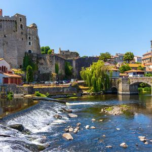 Un château médiéval en pierre et un pont en arc enjambent une rivière avec un petit barrage dans une ville historique sous un ciel bleu clair.