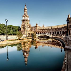 Un edificio decorato con un'alta torre si riflette nell'acqua calma di un canale, con un ponte che lo attraversa sotto un cielo azzurro limpido.