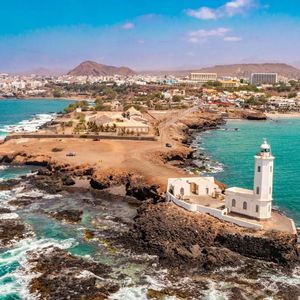 Vista aérea de un faro blanco en una península rocosa, con una ciudad costera y montañas al fondo bajo un cielo azul.