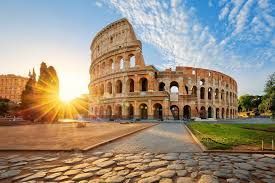 An ancient Roman amphitheater illuminated by a bright sunrise, with a cobblestone path in the foreground under a blue sky.