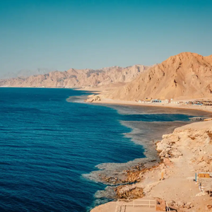 A panoramic view of a deep blue sea meeting a rugged, arid coastline with mountains under a clear sky.