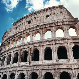 Un'inquadratura dal basso della struttura arcuata in pietra del Colosseo Romano contro un cielo azzurro con nuvole bianche.