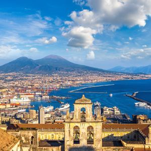 Una vista aerea di una città costiera e della sua baia, con un grande vulcano sullo sfondo, vista da dietro un campanile storico.