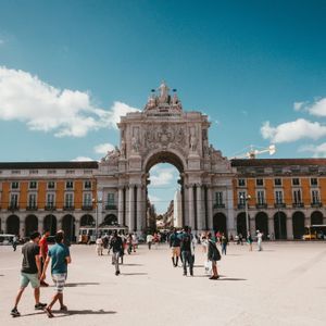 Persone che passeggiano in una grande piazza di fronte a un arco trionfale ornato e edifici gialli sotto un cielo azzurro e soleggiato.