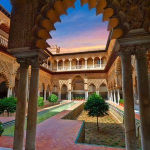 Un cortile in stile moresco riccamente decorato con una piscina centrale e giardini, visto attraverso un grande arco a conchiglia contro un cielo al tramonto.