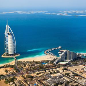 Vue aérienne d'un hôtel en forme de voile sur une petite île artificielle, relié par un pont à un littoral sablonneux et à une ville.