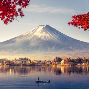 Une montagne enneigée se reflète sur un lac calme où une personne se tient dans une petite embarcation, encadrée par des feuilles d'automne rouges.