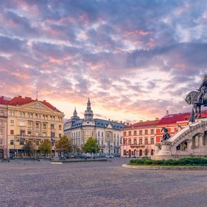 Una statua equestre si erge in una grande piazza acciottolata, circondata da edifici storici sotto un cielo colorato e nuvoloso al tramonto.