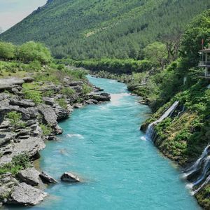Ein türkiser Fluss schlängelt sich durch eine felsige Schlucht, wo kleine Wasserfälle einen üppigen Berghang neben einem Gebäude hinabstürzen.