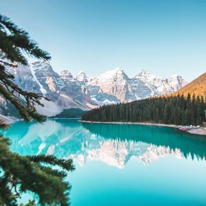 Montagnes enneigées et forêt de pins dense se reflétant dans l'eau calme et turquoise d'un lac sous un ciel clair.