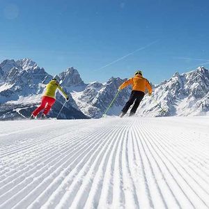 Zwei Skifahrer in bunten Jacken fahren eine präparierte Skipiste hinab mit Panoramablick auf eine Bergkette unter klarem Himmel.