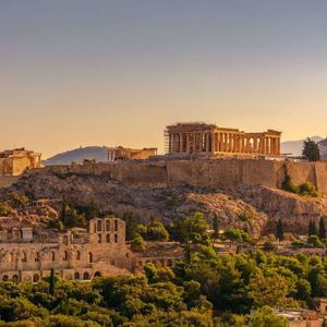 Antichi templi e rovine greche si ergono su una collina rocciosa che domina una fitta foresta durante un tramonto dorato.
