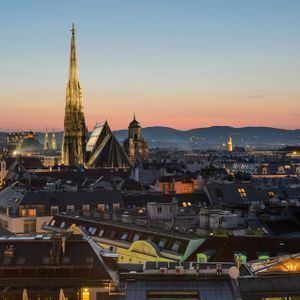An illuminated cathedral spire rises above city rooftops at twilight, with distant mountains silhouetted against a colorful sky.