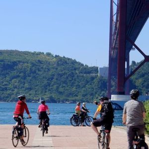 Un viaggio di gruppo WeRoad in bicicletta su un percorso lungomare con un grande ponte rosso e un monumento su una collina sullo sfondo.