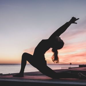 Una silhouette di una persona in posa yoga su un tappetino su un molo in riva al mare al tramonto.