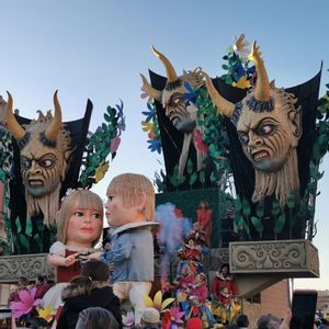 Un elaborato carro di carnevale, caratterizzato da grandi teste di demoni cornuti e due bambole giganti, attraversa una strada affollata.