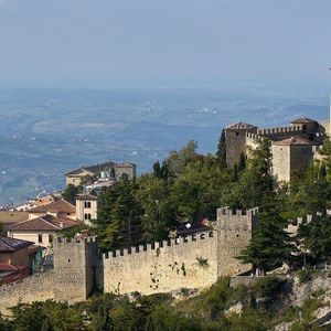Un castello medievale in pietra con mura fortificate è arroccato su una montagna rocciosa e alberata, che domina una valle di dolci colline.