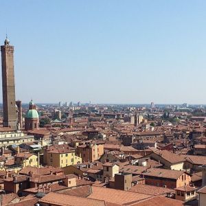 Un paesaggio urbano di tetti in terracotta, con un'alta torre di mattoni e una chiesa a cupola che si ergono contro un cielo azzurro chiaro.