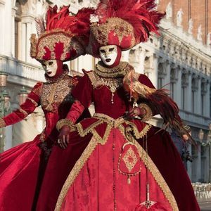 Due figure in elaborati costumi di velluto rosso, copricapi piumati e maschere bianche posano in un'ampia piazza pubblica illuminata dal sole.