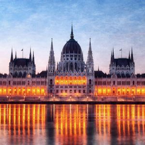 A large, illuminated government building with spires and a dome reflects in a river at twilight.