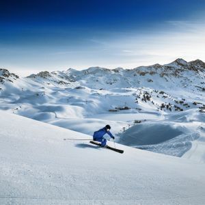 Uno sciatore in tuta blu scende un vasto pendio montano innevato sotto un cielo azzurro limpido.