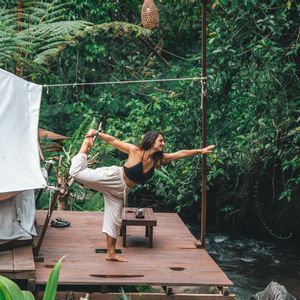 Una donna esegue una posa yoga in piedi su una piattaforma di legno accanto a un fiume, circondata da una fitta vegetazione verdeggiante della giungla.