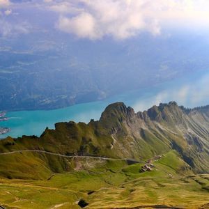 Vue plongeante sur des montagnes verdoyantes avec une route sinueuse, dominant un lac turquoise et une vallée abritant une ville.