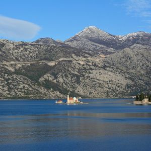Due piccole isole, una con una chiesa e l'altra con alberi, si trovano su una tranquilla baia blu davanti a una grande catena montuosa rocciosa.