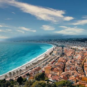 Vue aérienne d'une ville côtière aux toits orangés, s'étirant le long d'une plage bordée par des eaux turquoise, sous un ciel bleu parsemé de nuages.