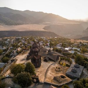 Un antico monastero in pietra e un villaggio circostante su una collina soleggiata con montagne sfocate sullo sfondo.