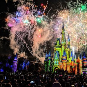 Colorful fireworks explode over a brightly illuminated fairytale castle as a large crowd watches the show at night.